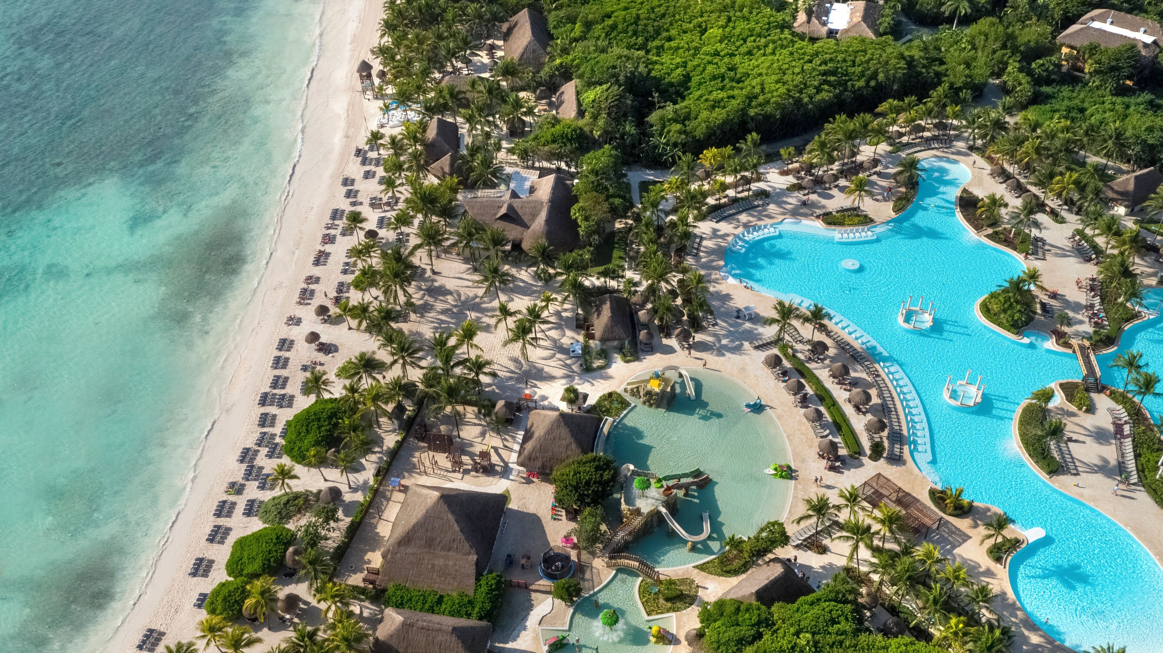 Vista aérea de um resort com praia, palmeiras e uma grande piscina com toboágua.