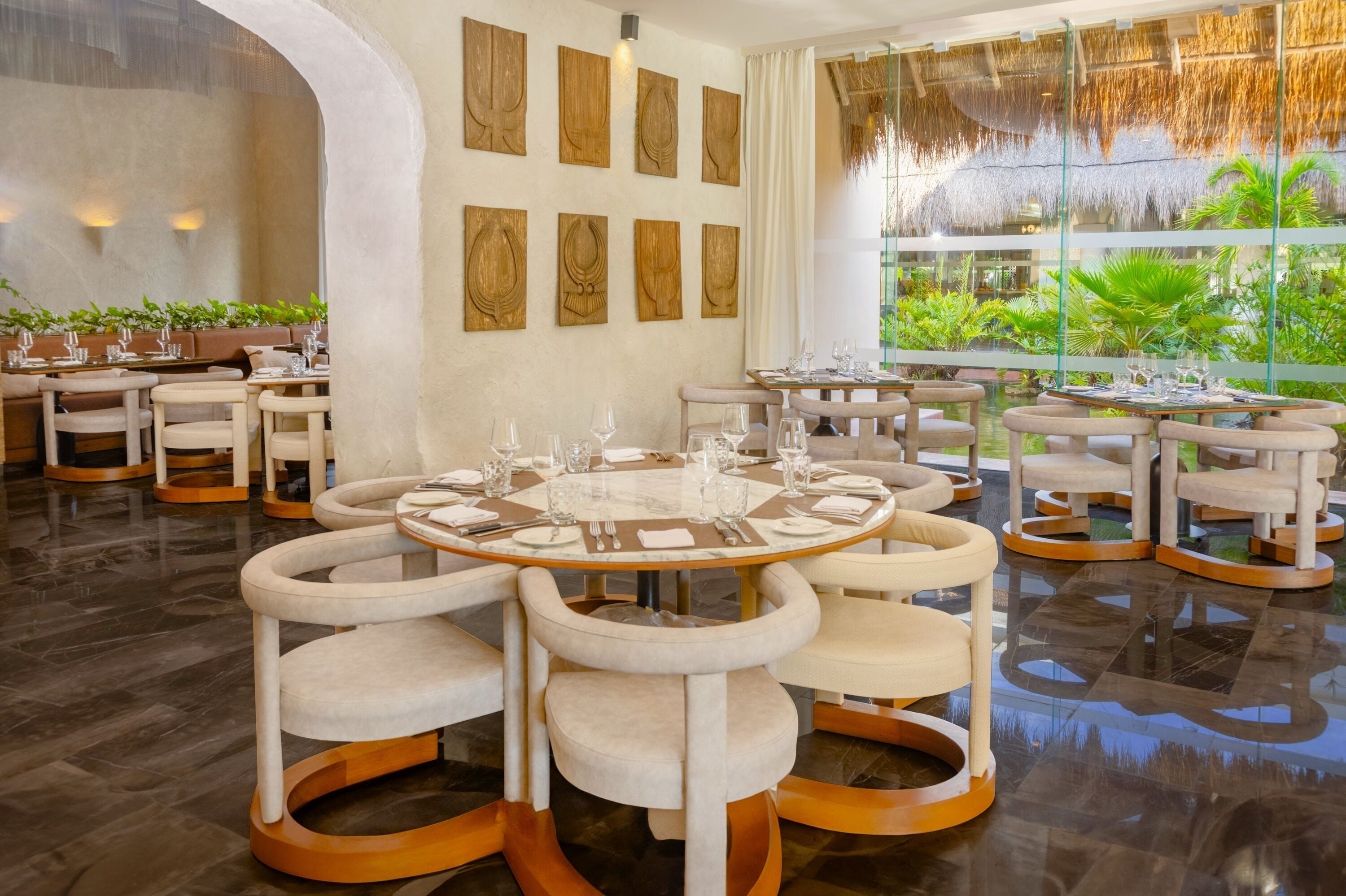 Un comedor moderno con mesas redondas y sillas acolchadas, rodeado por un muro de cristal con vistas a la vegetación.