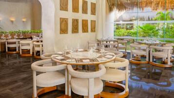 Un comedor moderno con mesas redondas y sillas acolchadas, rodeado por un muro de cristal con vistas a la vegetación.