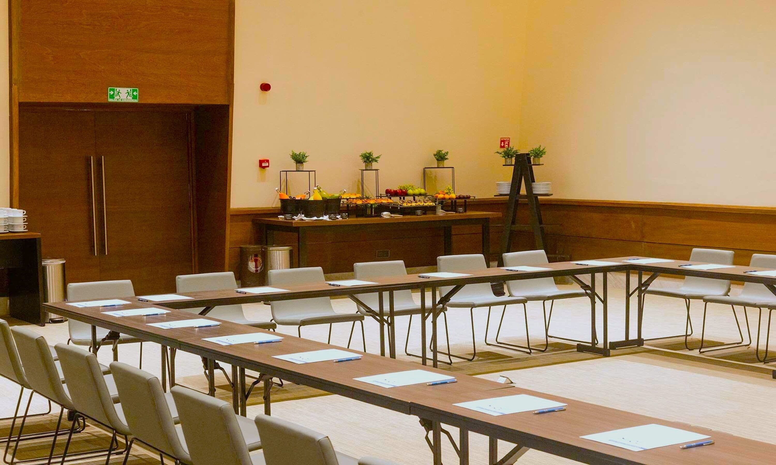 A conference room with long tables, chairs, and a buffet table with fruits and vegetables.