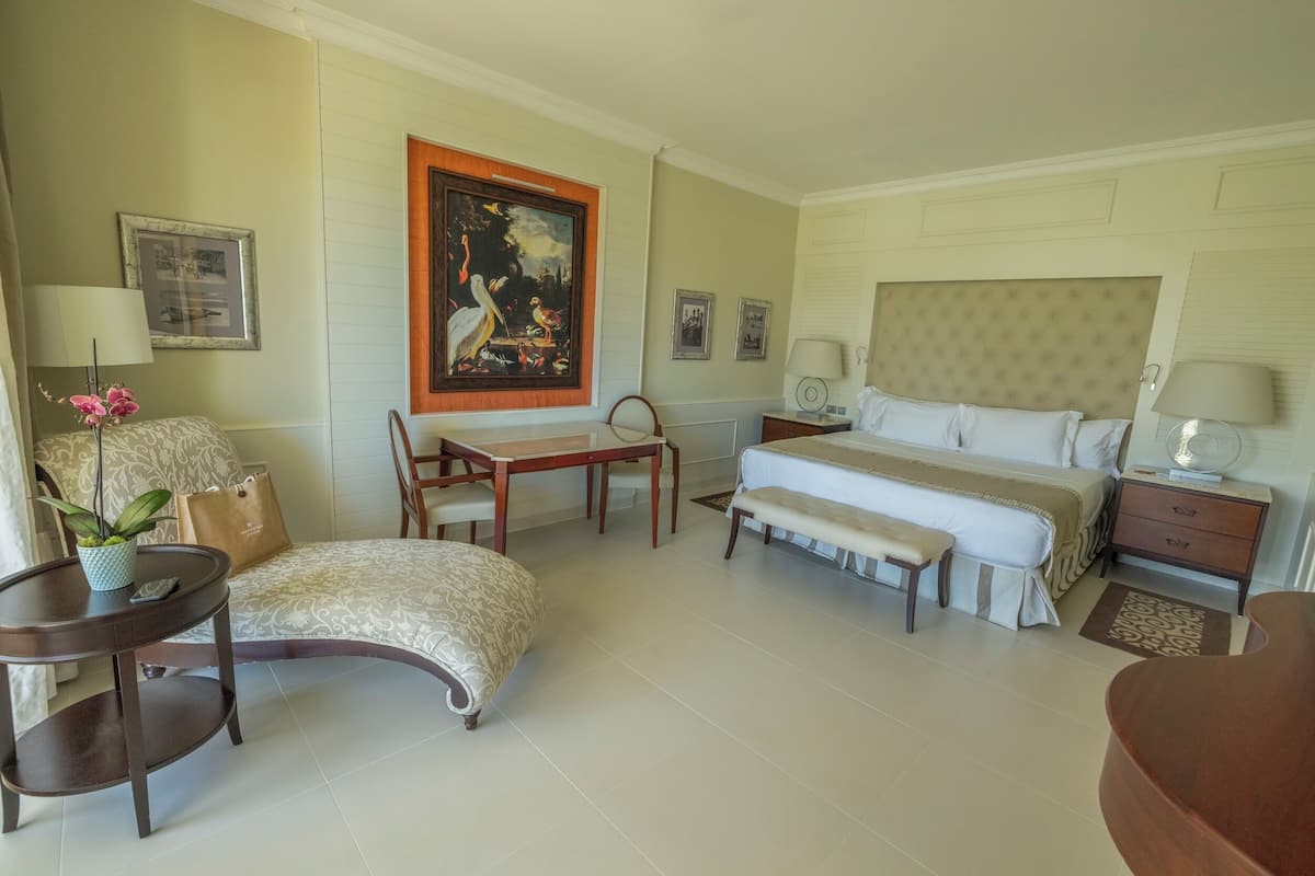 A hotel room with a bed, bedside tables, a desk, a chair, a small table with a plant, and a bench.