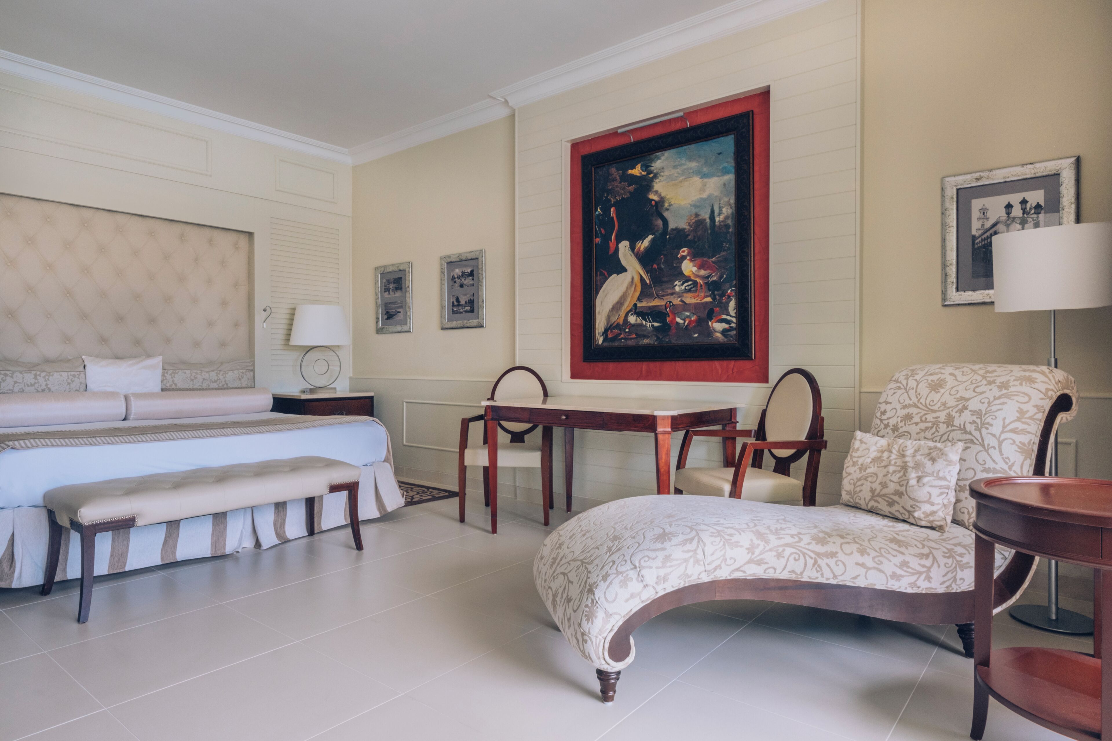 A hotel room with a bed, a bench, a table, a chair, and a painting on the wall.
