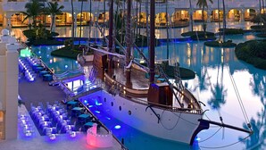 A large indoor swimming pool with a ship-shaped structure, surrounded by palm trees and a hotel building in the background. - JOIA Bávaro by Iberostar - Adults Only - All Inclusive (Punta Cana)