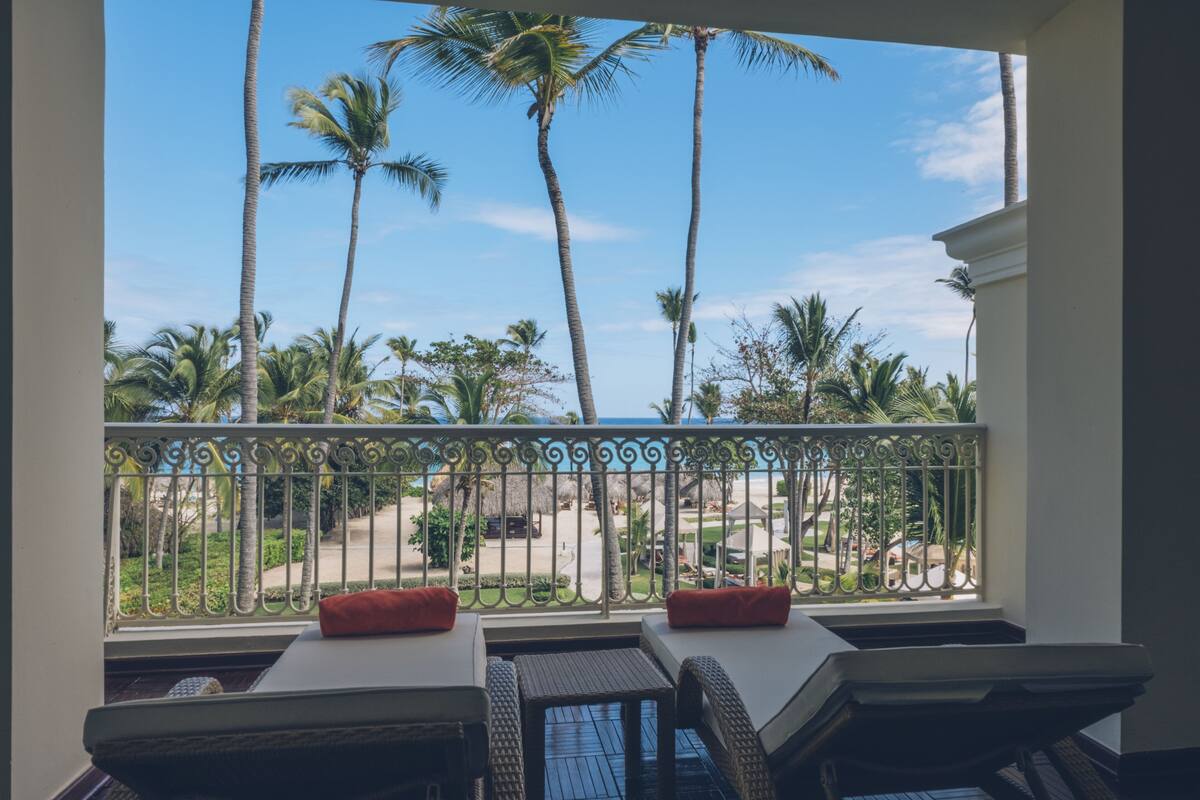 Ein Balkon mit Korbmöbeln und roten Kissen mit Blick auf einen Strand, der mit Palmen und einem klaren Himmel gesäumt ist.