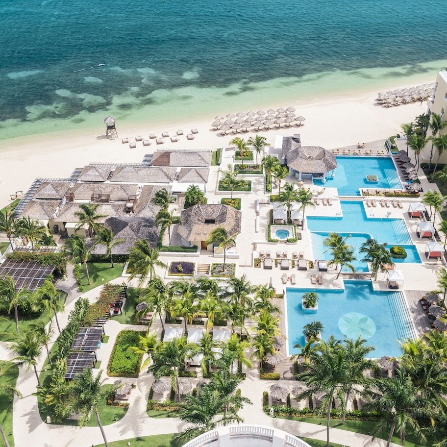 An aerial view of a resort with multiple swimming pools, a beach, and palm trees.