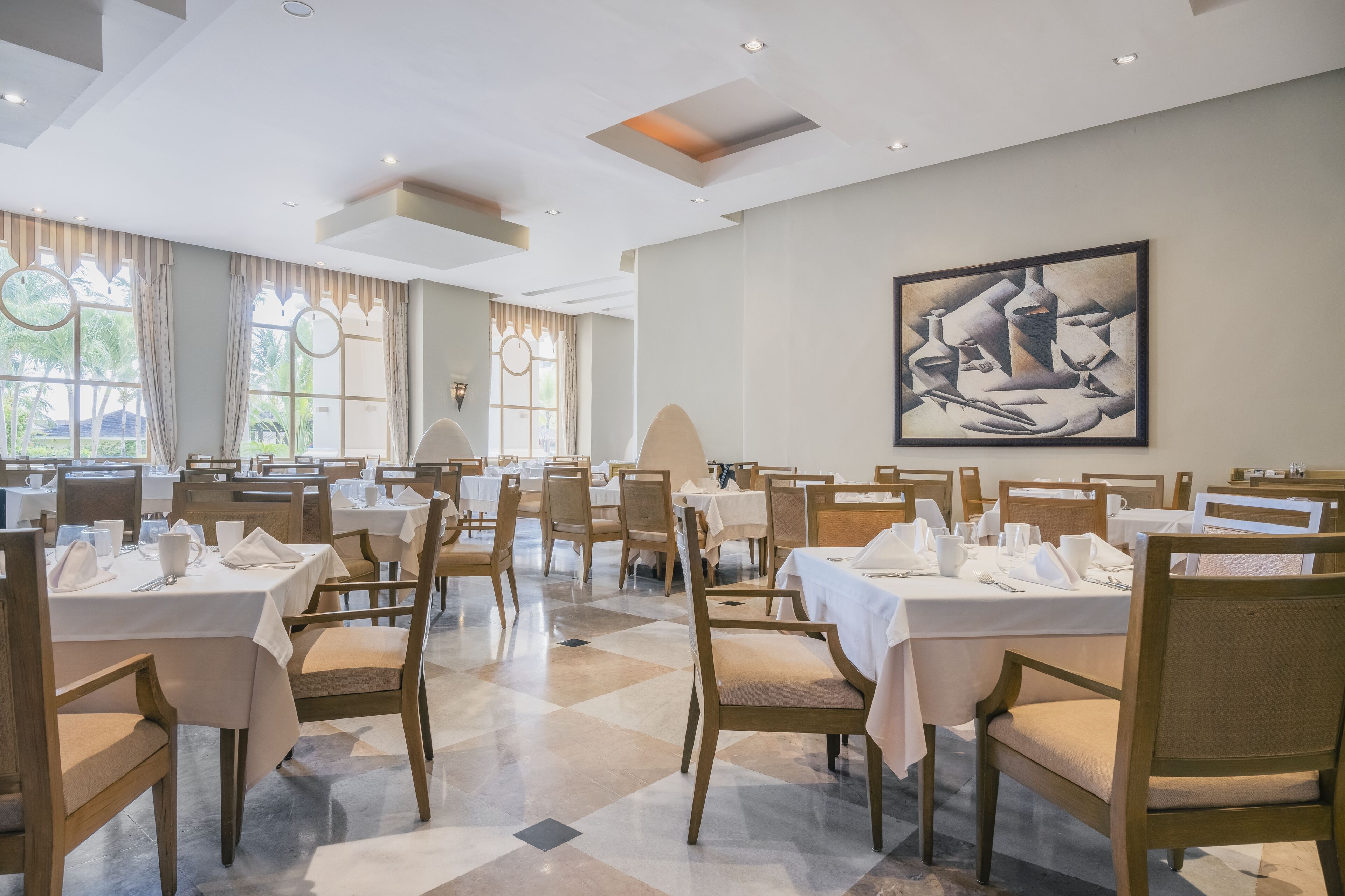A dining area with tables set for service, wooden chairs, and a large abstract painting on the wall.