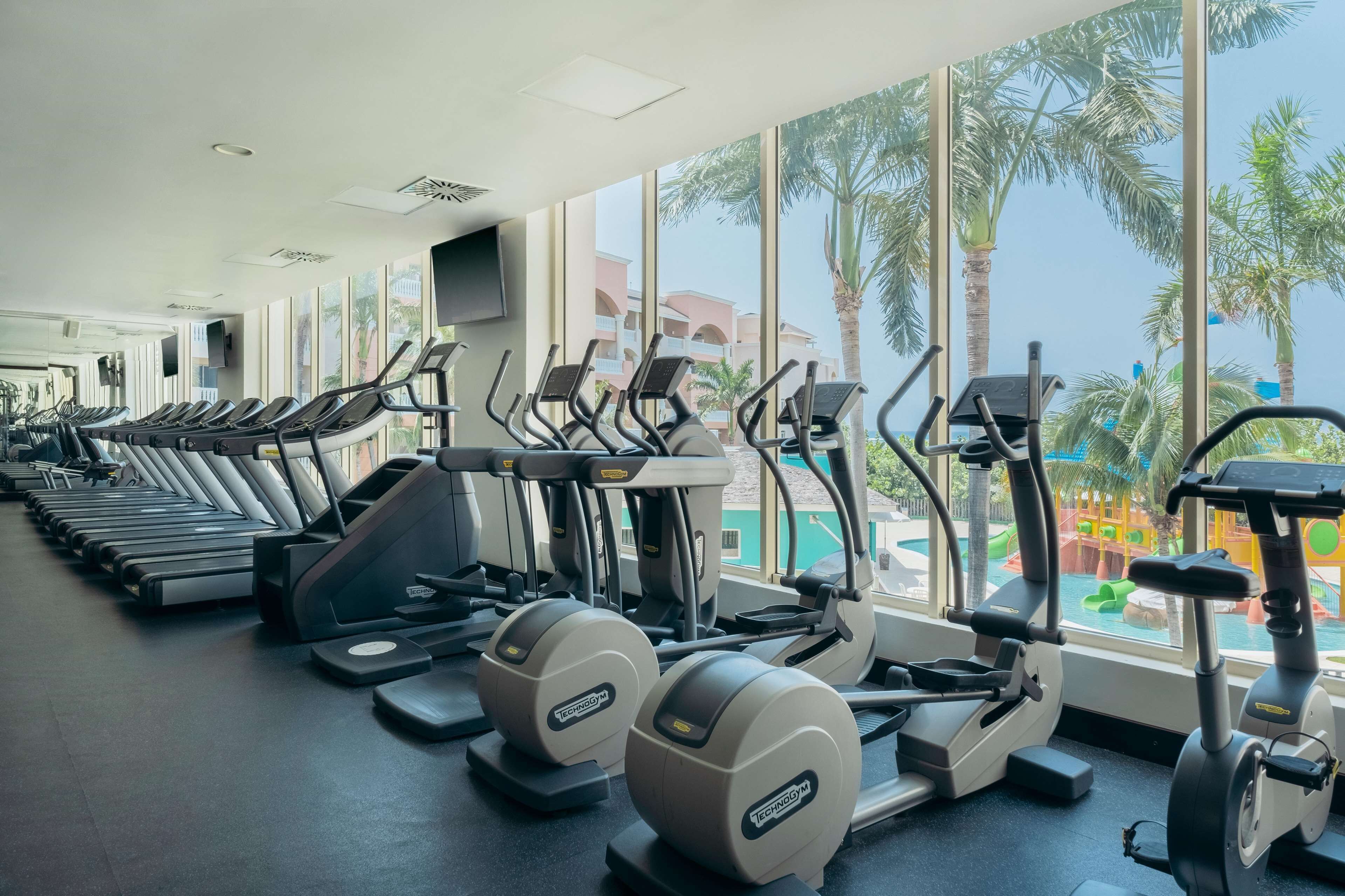 A gym with treadmills, elliptical machines, and stationary bikes, large windows with a view of palm trees and a pool.