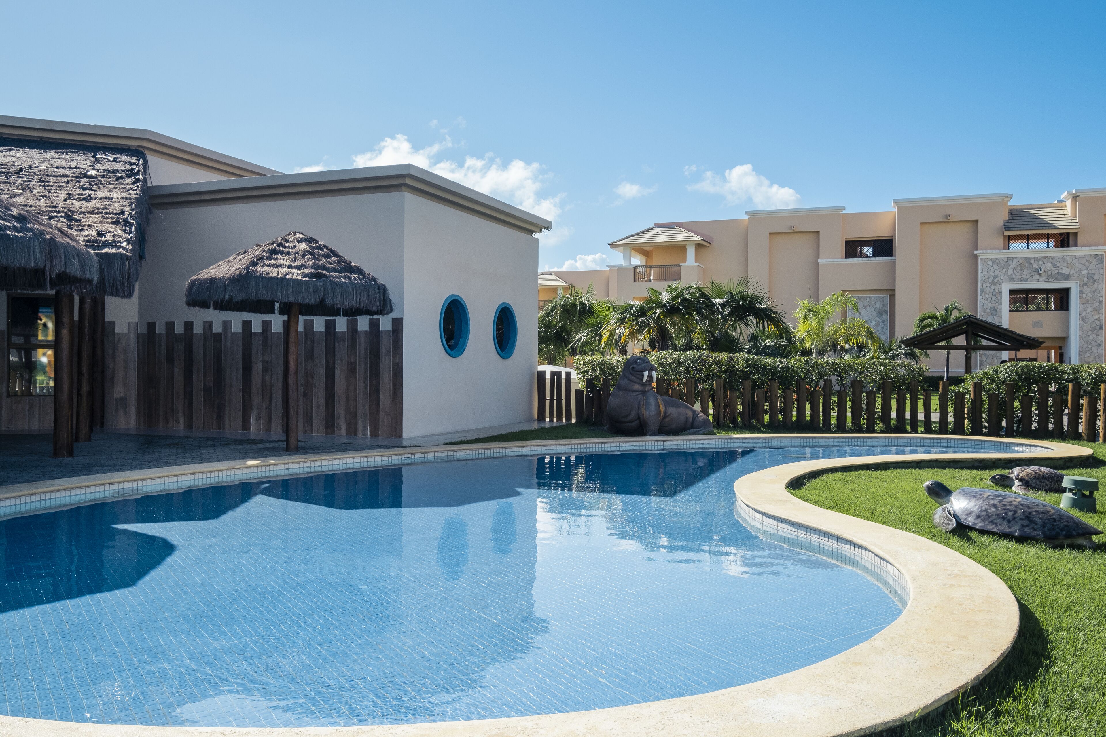 Uma piscina com borda curva, uma estrutura de telhado de palha e uma cerca de madeira.
