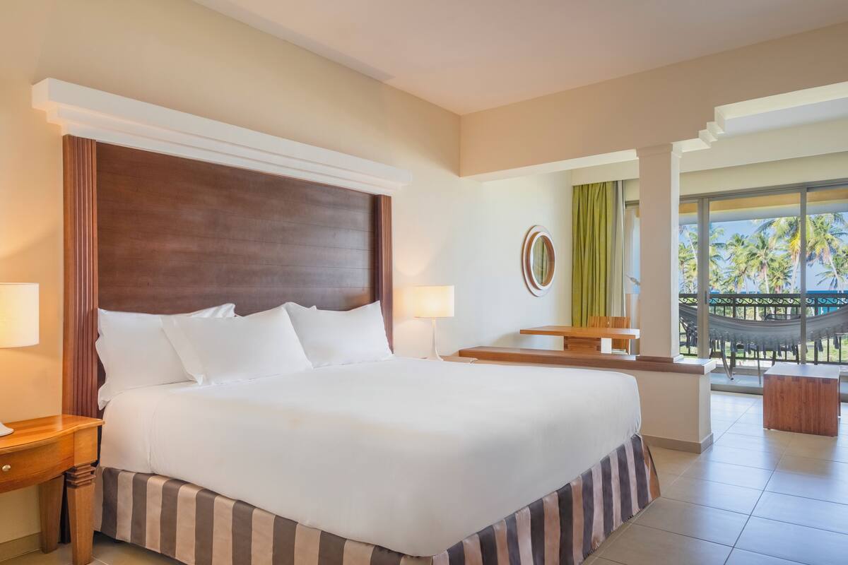 Ein Hotelzimmer mit einem großen Bett, zwei Nachttischen, einem kleinen Schreibtisch, einem Sessel und einem Balkon mit einer Hängematte.