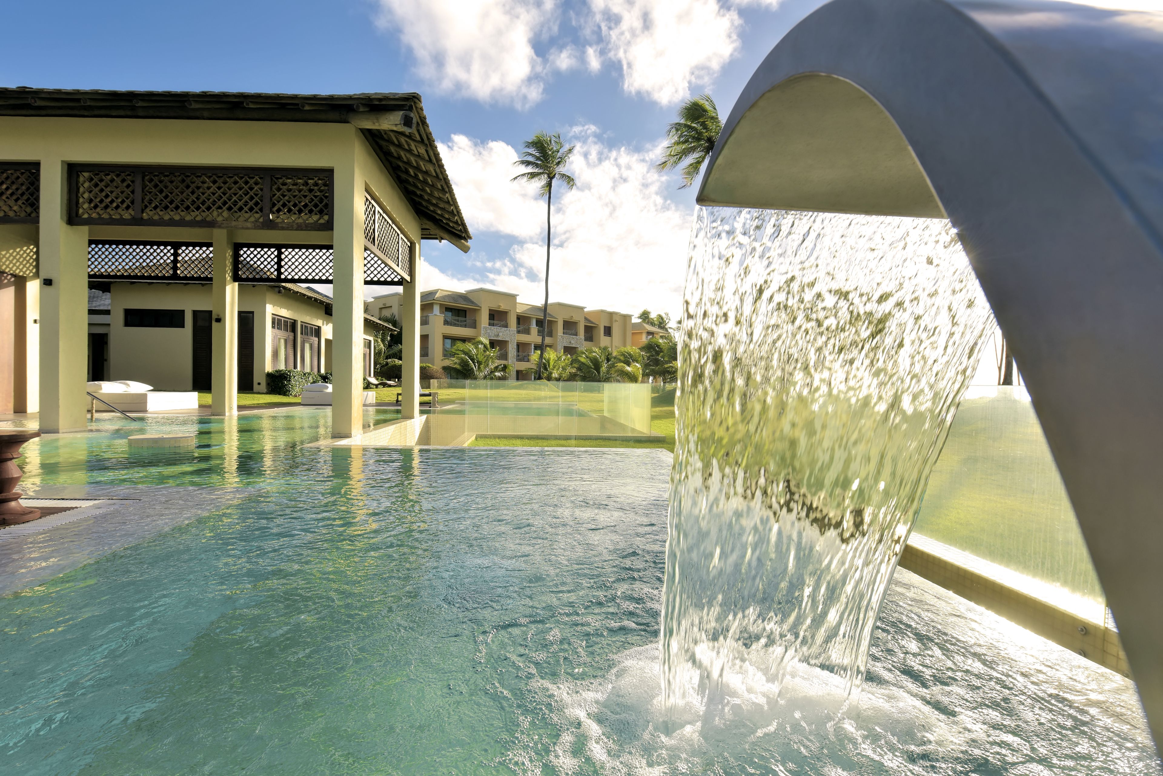 Ein Pool mit Wasserspiel, ein überdachter Sitzbereich und im Hintergrund ein Gebäude mit Balkonen.