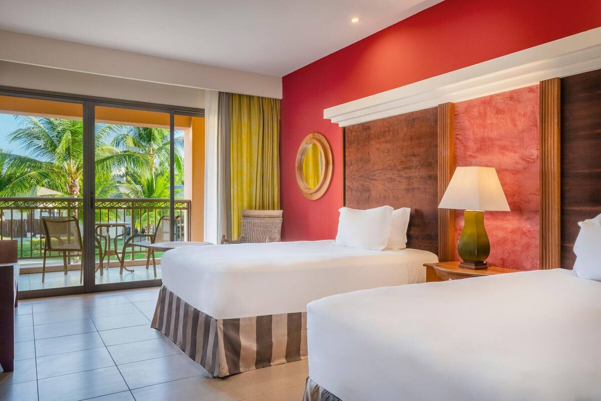 Ein Hotelzimmer mit zwei Betten, einem Balkon mit Blick auf Palmen und einer Wand mit Holzverkleidung.