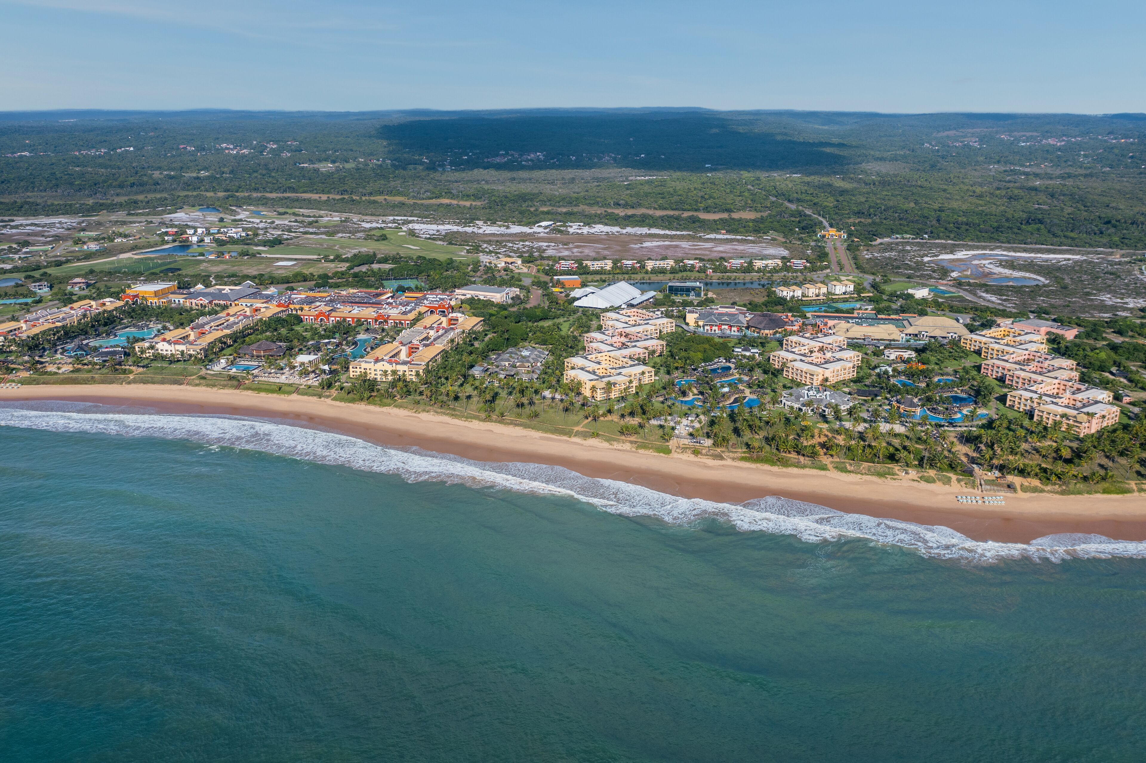 Vista aérea de un complejo turístico costero con villas frente a la playa, una playa de arena y colinas con vegetación al fondo.