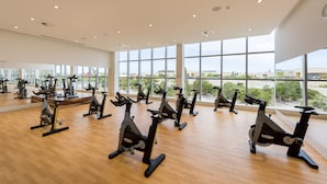 Een ruime sportschool met meerdere vaststaande fietsen in rijen, grote ramen met uitzicht naar buiten, en een houten vloer.