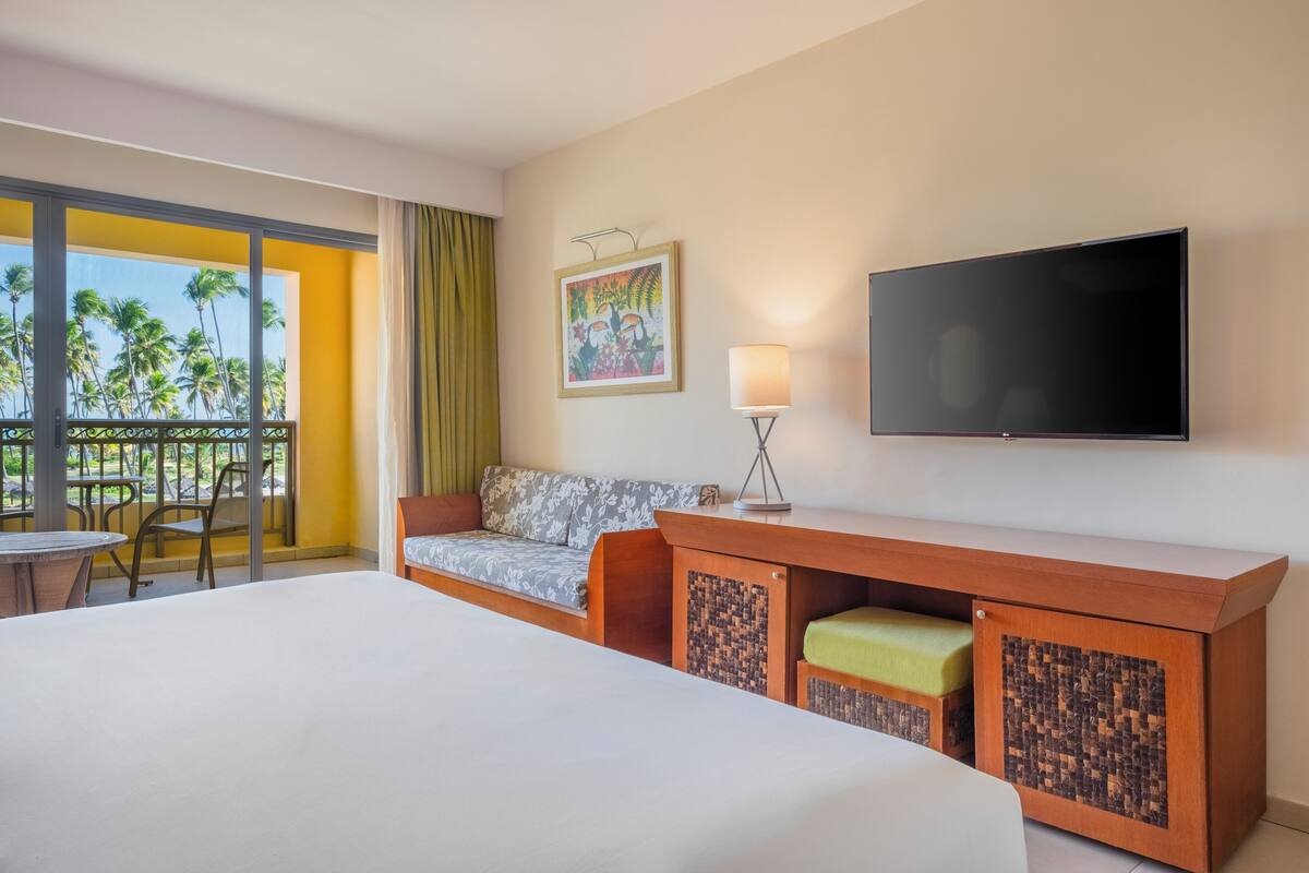 Ein Hotelzimmer mit einem großen Bett, einer Couch, einem Schreibtisch, einem Fernseher und einem Balkon mit Blick auf Palmen.