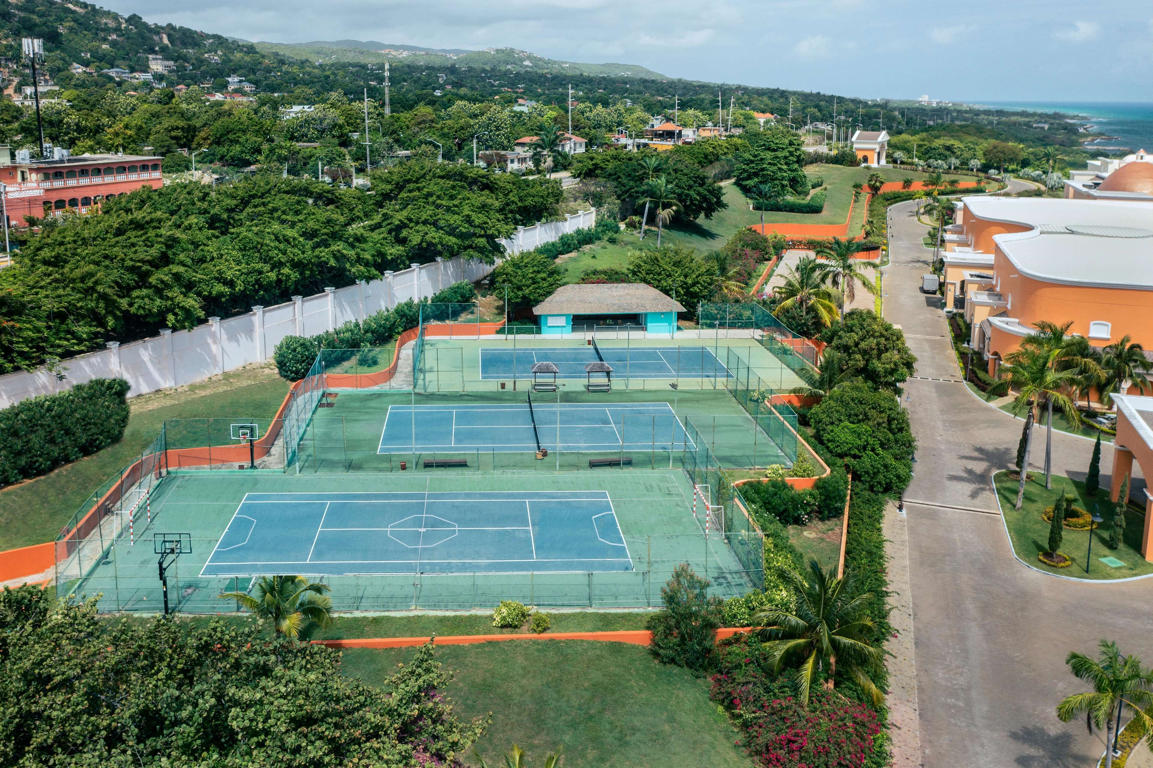 Eine Vogelperspektive zeigt einen Tennisplatzkomplex, umgeben von üppiger Vegetation und Wohngebäuden.