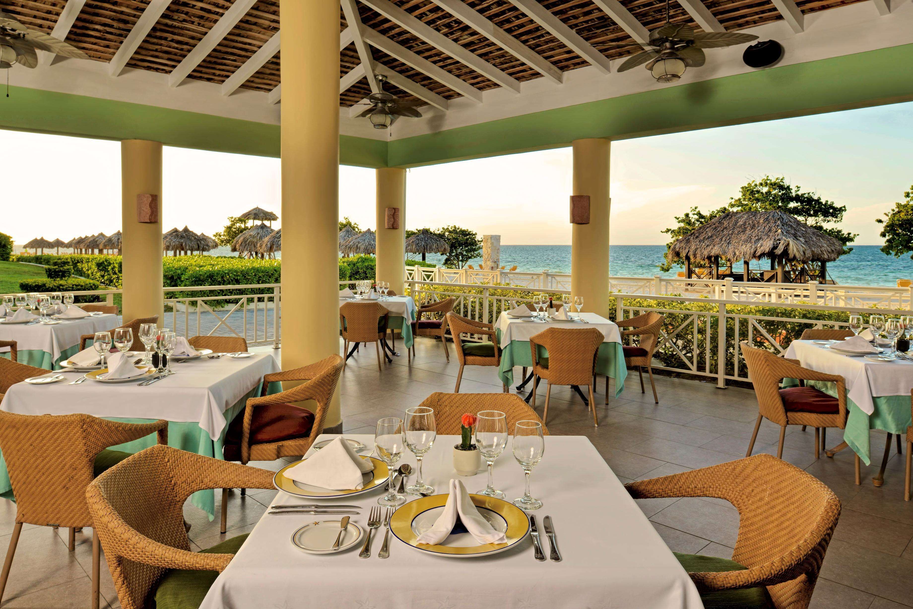 A restaurant with tables set for dining, overlooking a beach with thatched-roof structures.