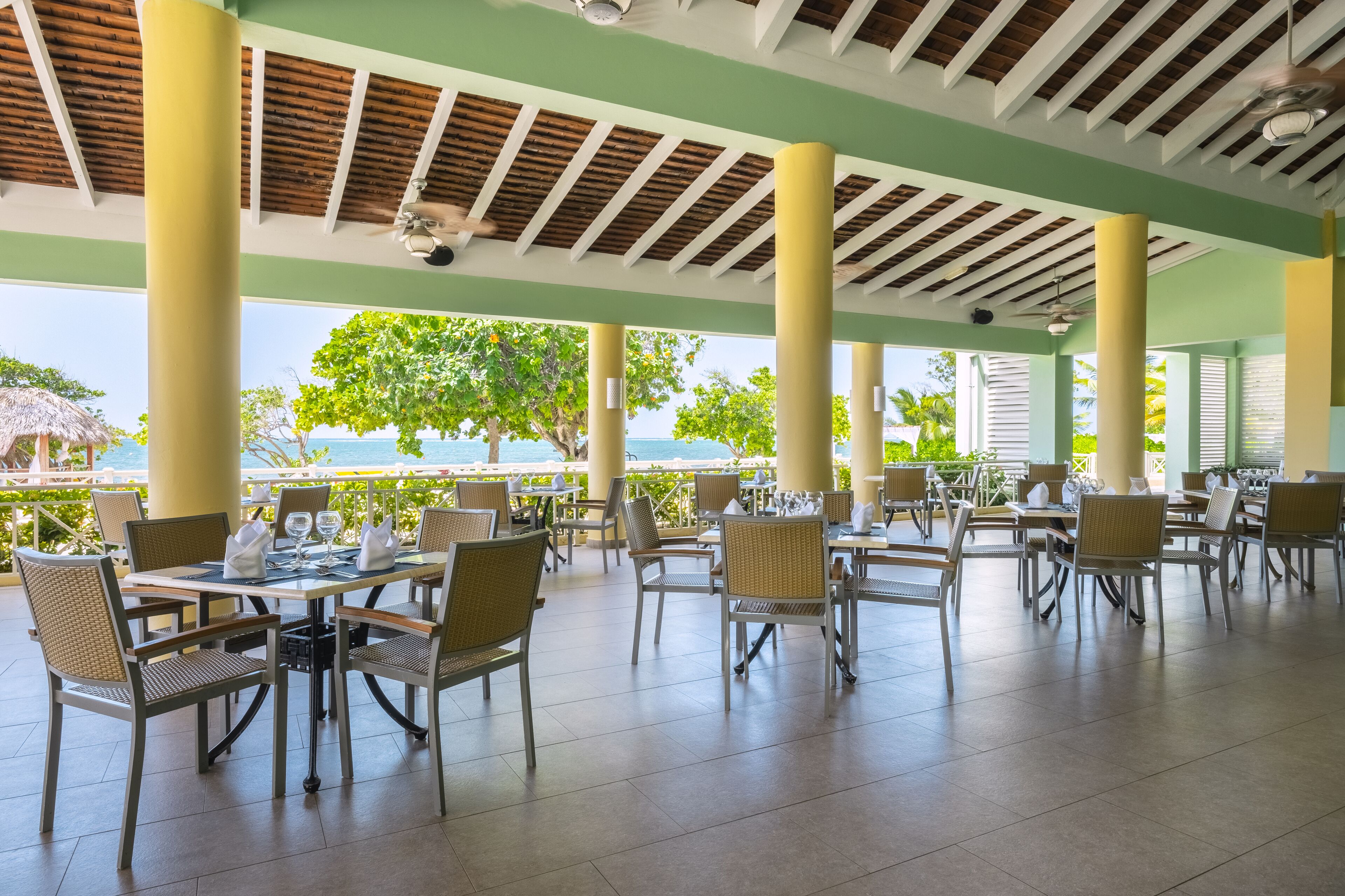 An outdoor dining area with tables and chairs, a view of the sea, and a covered roof structure.