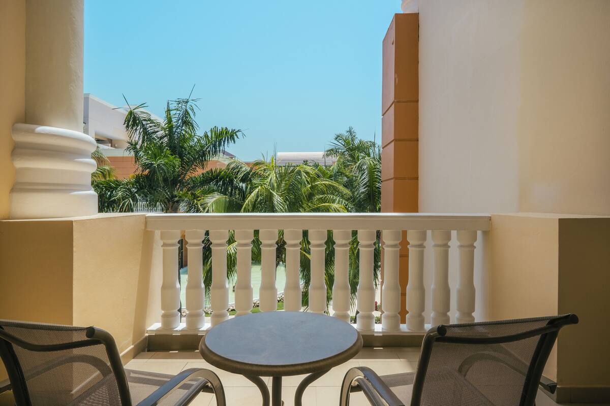 A balcony with a round table and two chairs, a white balustrade, and a view of palm trees and buildings.