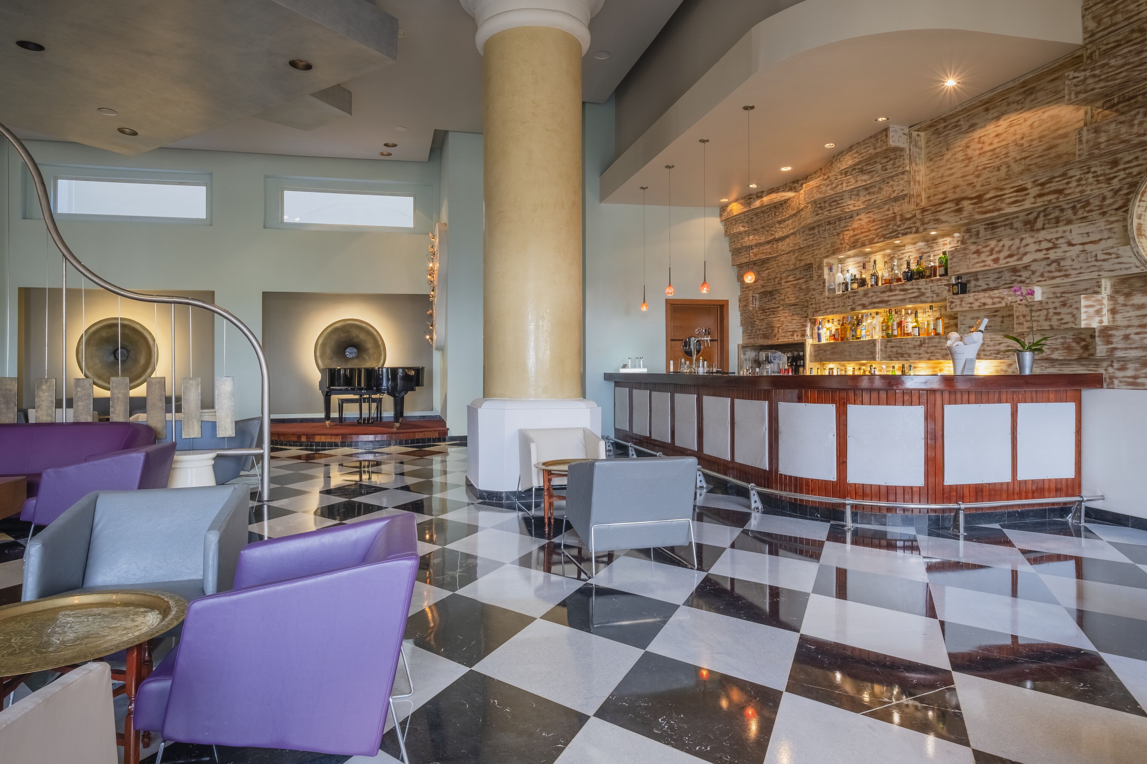 A lobby with checkered flooring, purple and gray seating, a central column, a bar area with a stone wall, and a staircase with circular light fixtures.