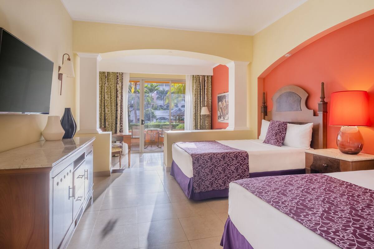 A hotel room with two beds, a TV, a desk, and a view of the outdoors.