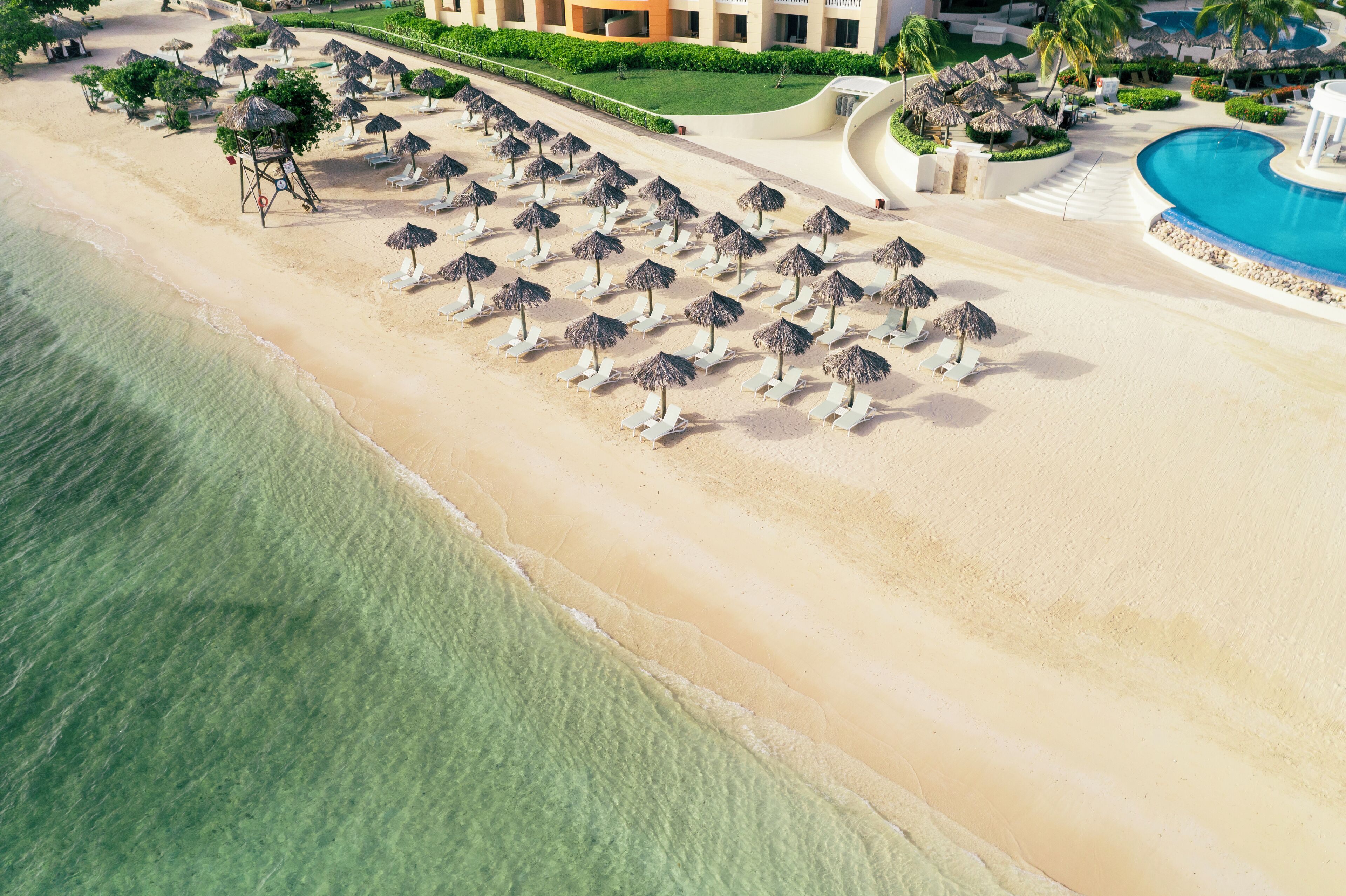 An aerial view of a beach with rows of sun loungers and umbrellas, a swimming pool, and a resort building.