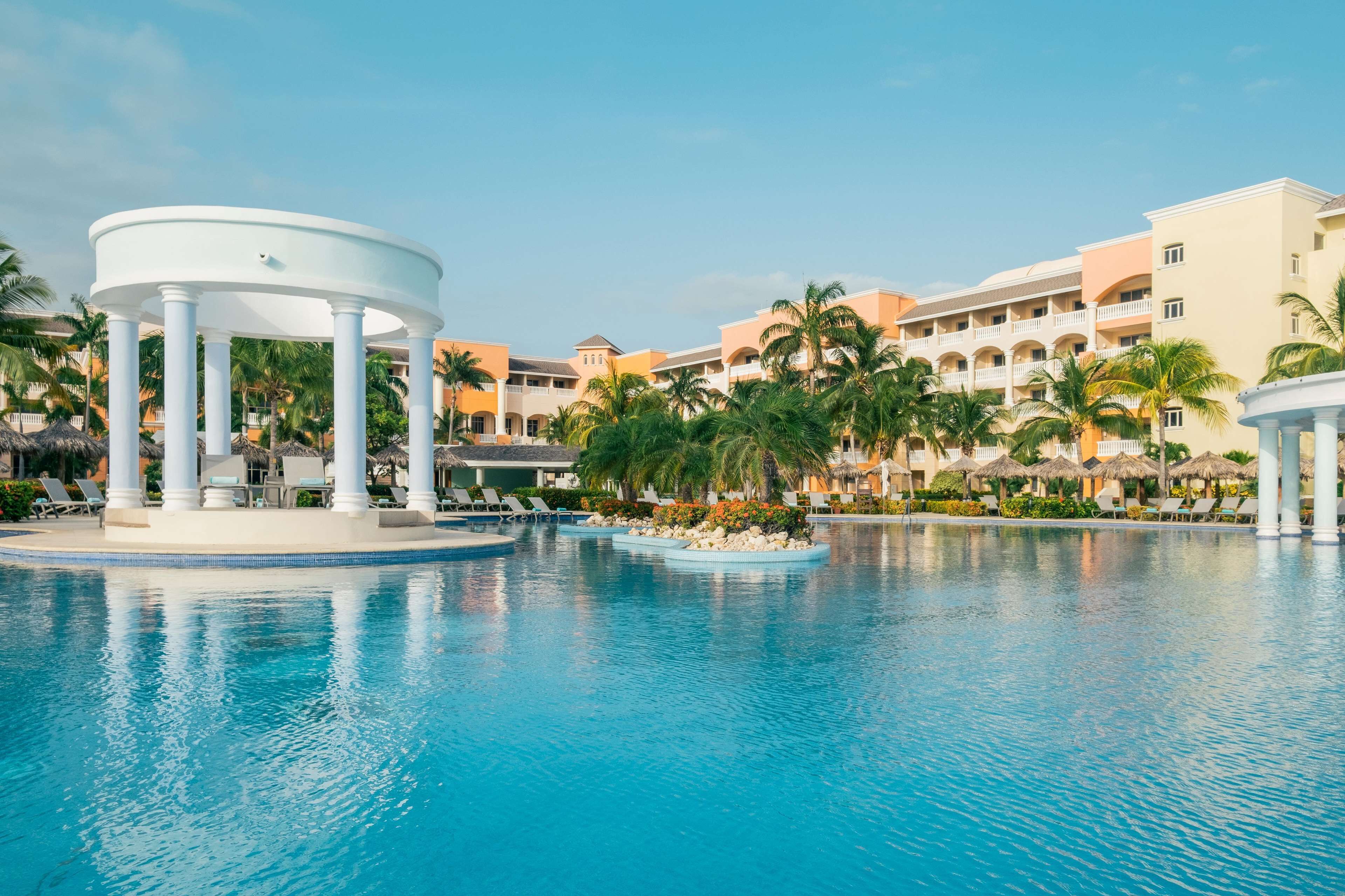 A resort pool area with a central circular structure, palm trees, and resort buildings in the background.