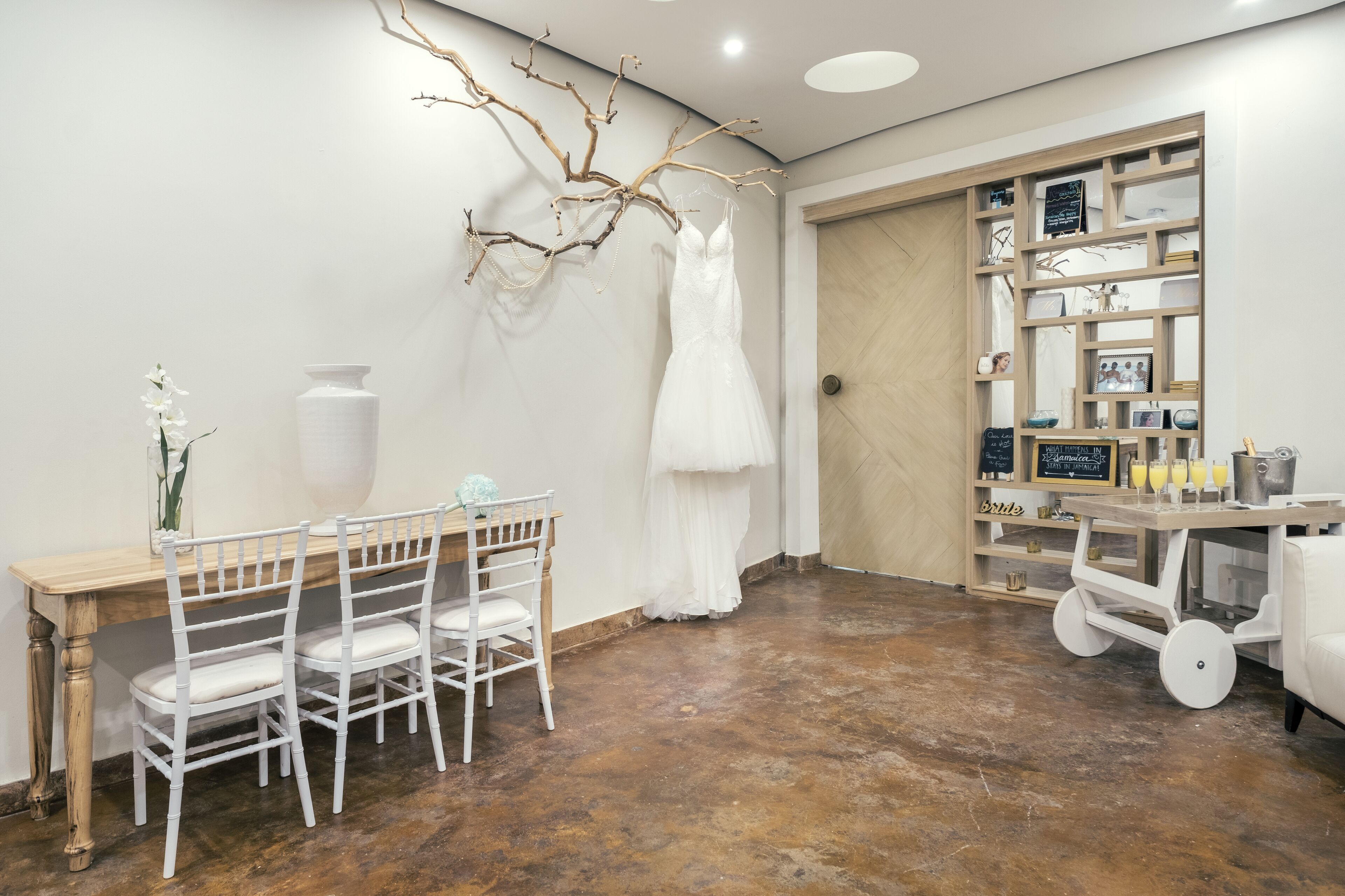 A room with a wooden table and chairs, a white dress on a branch, and a shelf with framed photos and a cart with drinks.