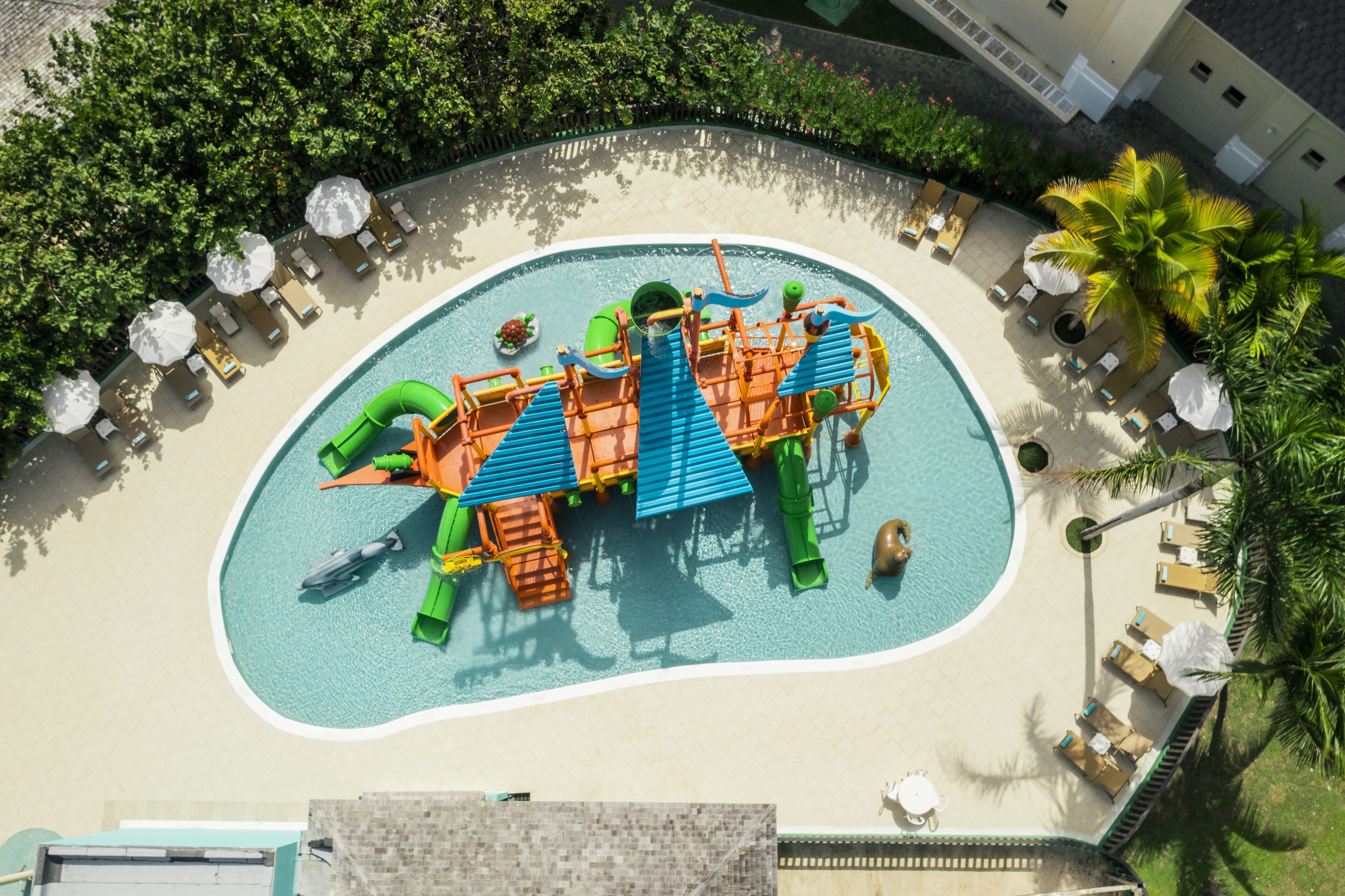 An aerial view of a resort's swimming pool area with a water slide, lounge chairs, and palm trees.