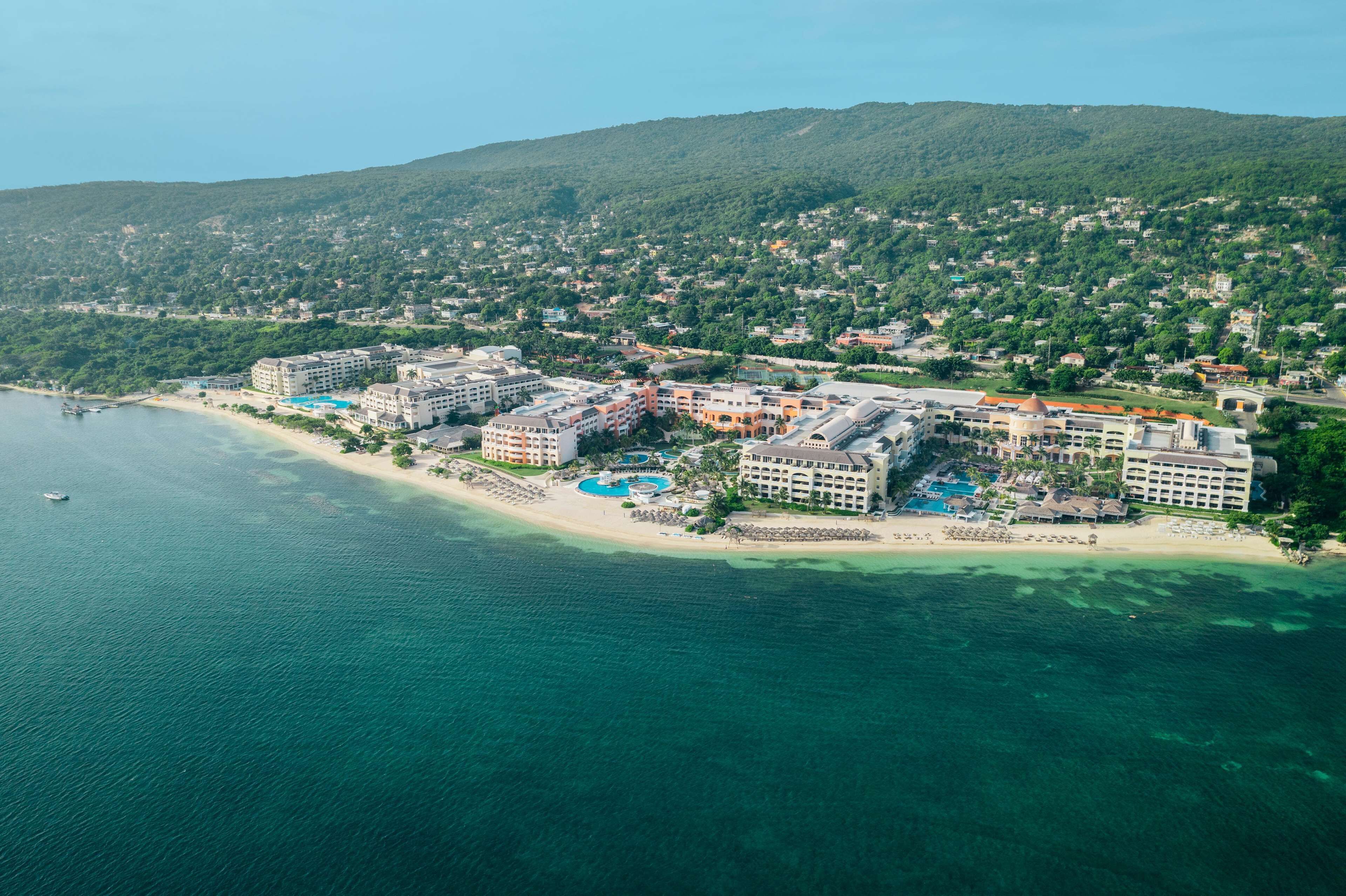 An aerial view of a coastal resort with multiple buildings, a beach, and a mountainous backdrop.