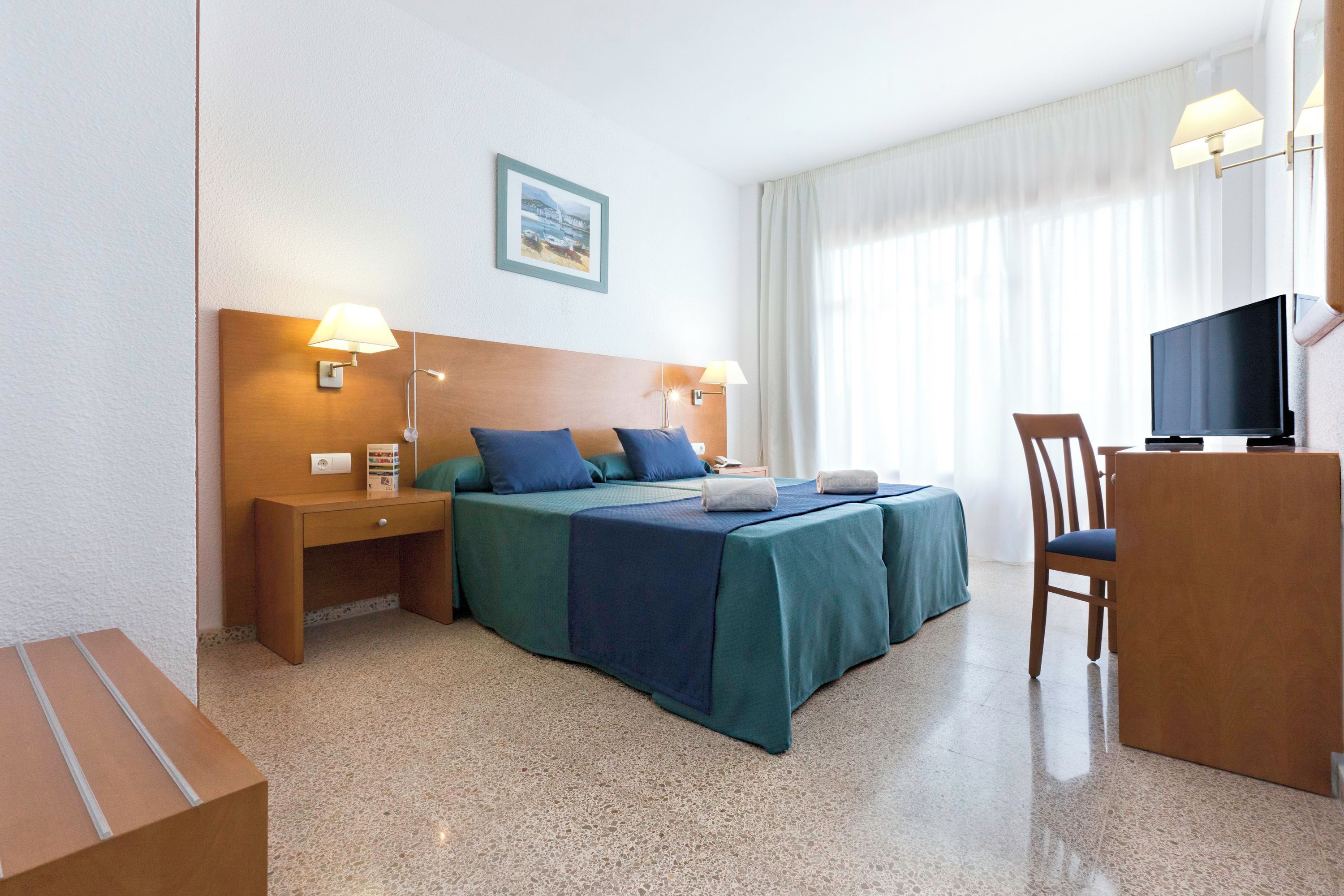 A hotel room with a bed, bedside tables, a desk with a chair, a television, and a window with curtains.