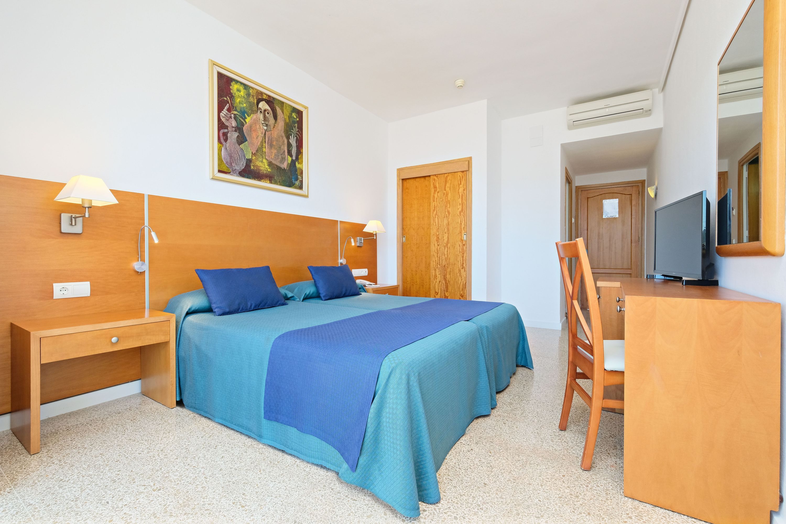 Habitación de hotel con cama, mesitas de noche, un escritorio, una silla, televisor y un cuadro en la pared.