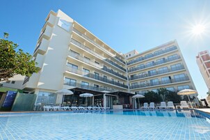 Un hotel de varios pisos con piscina, asientos al aire libre y un cielo azul despejado.