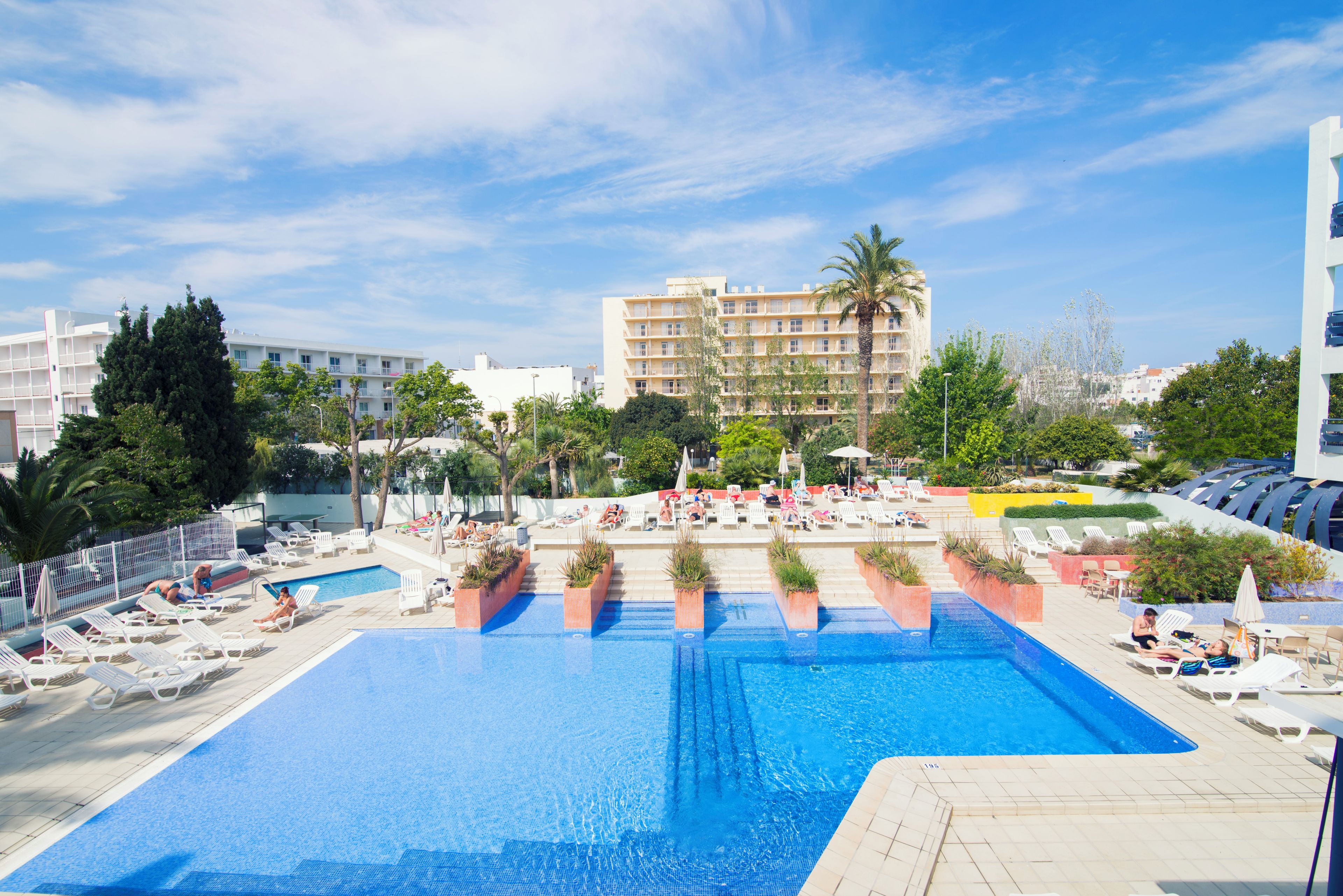 Una gran piscina al aire libre con sillones y tumbonas, rodeada de palmeras y edificios.