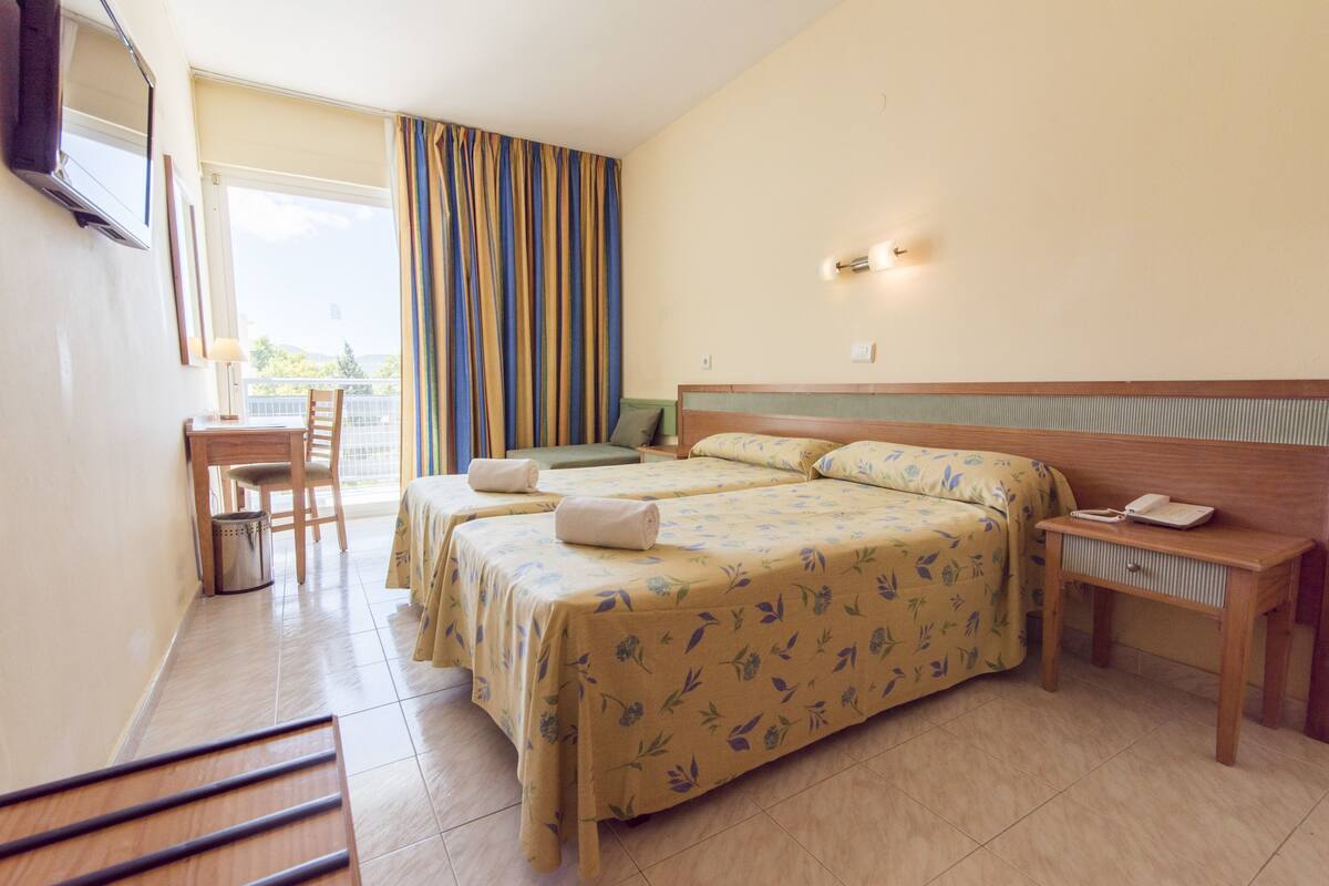 Habitación de hotel con dos camas, un escritorio, una silla, una mesita y una ventana con cortinas.