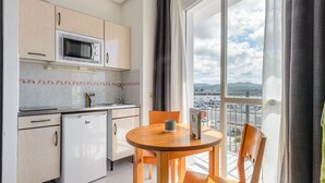 Eine kleine Küche mit Mikrowelle, Kühlschrank und Spüle. Ein Holztisch mit Stühlen steht in der Nähe eines großen Fensters mit Blick auf einen Hafen und ferne Berge.