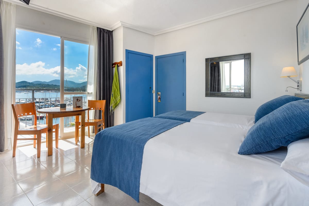 Une chambre d’hôtel avec un grand lit, une petite table avec des chaises, un balcon donnant sur la mer et deux portes bleues.