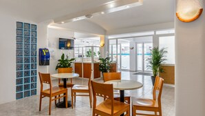 A modern hotel lobby with round tables and wooden chairs, a reception desk, and potted plants.