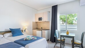Ein Hotelzimmer mit zwei Betten, einer Küchenzeile, einem kleinen Esstisch und einem Fenster mit Blick auf Bäume.