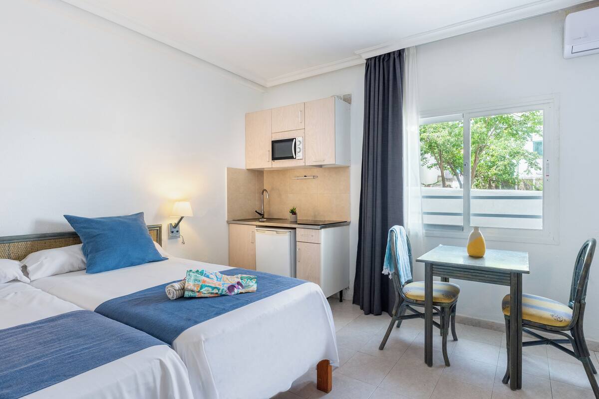 Une chambre d’hôtel avec deux lits, une kitchenette, une petite table à manger et une fenêtre donnant sur des arbres.