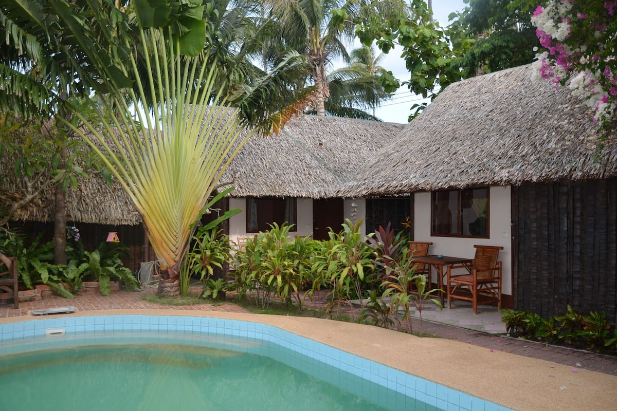 A poolside area with a thatched-roof building, surrounded by tropical plants and a palm tree.