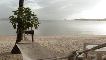 A hammock tied to a tree on a sandy beach with a view of calm water and distant land.
