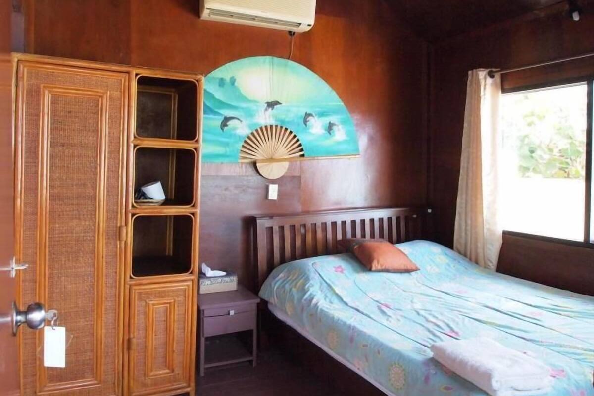 A room with a wooden wardrobe, a bed with a blue bedspread, a wall-mounted fan, and a window with curtains.