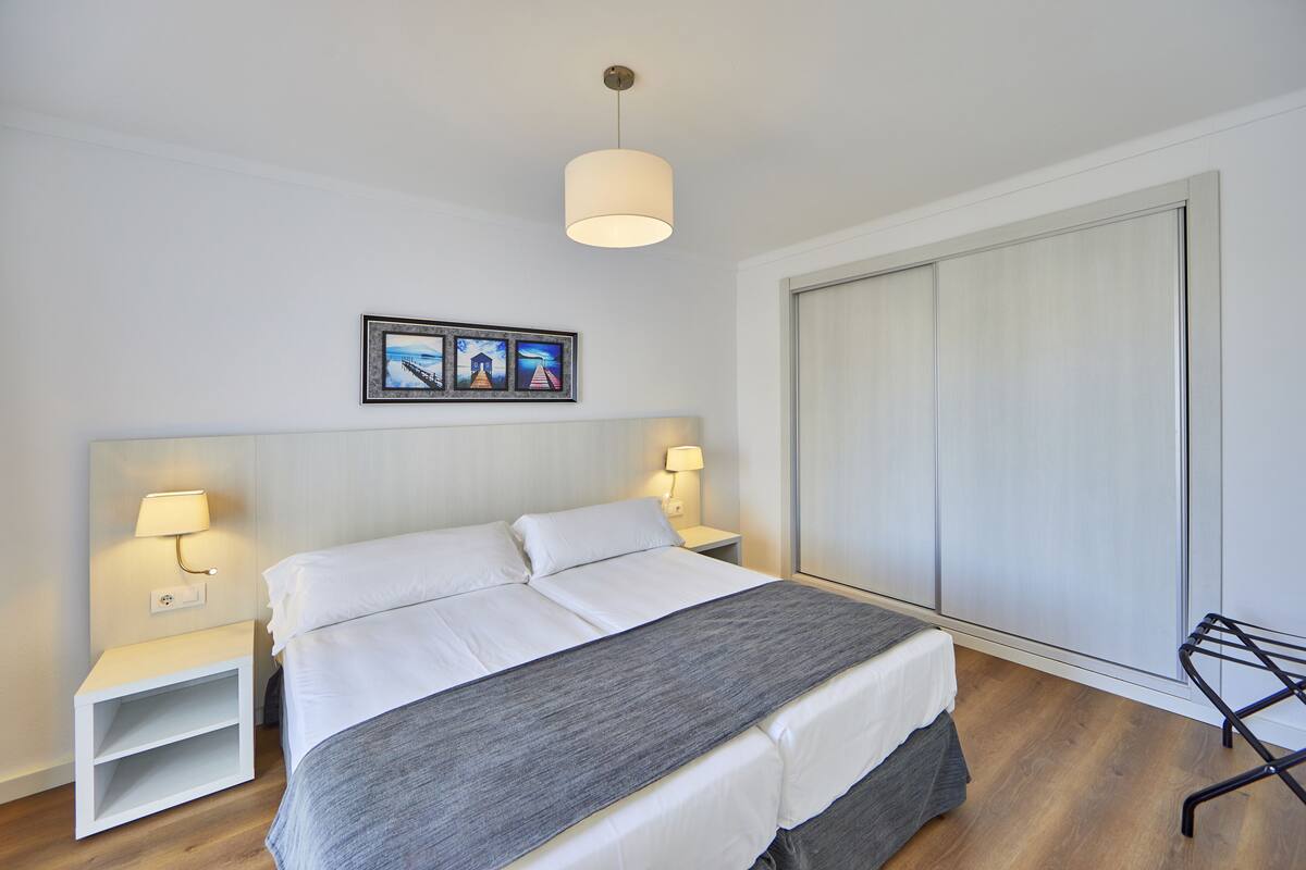 Una cama bien hecha con sábanas blancas, un plaid gris y dos lámparas de noche. Hay un cuadro enmarcado sobre la cama y una ventana grande con persianas a la derecha.