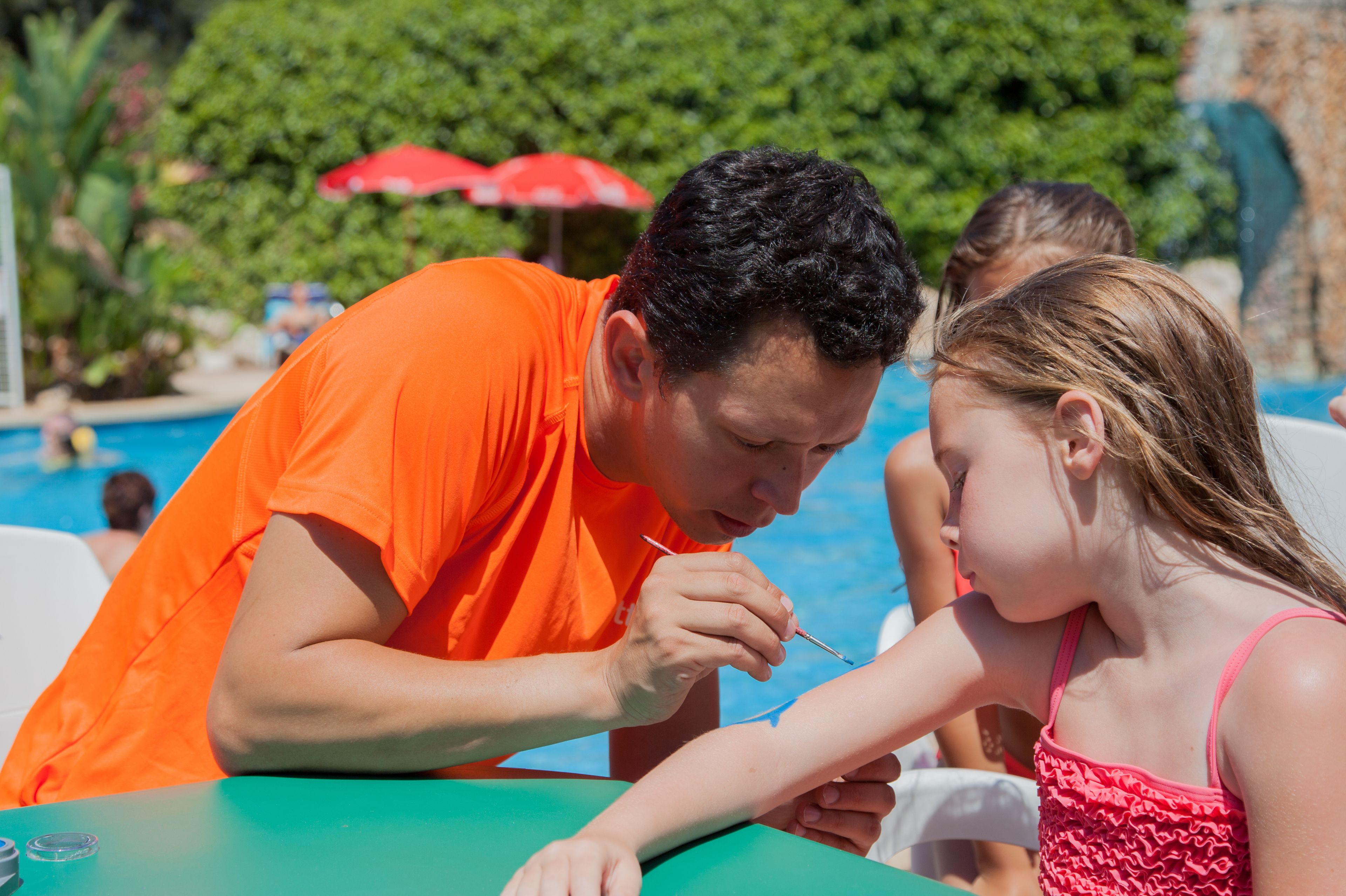 A man in an orange shirt applying sunscreen to a child at a poolside table.