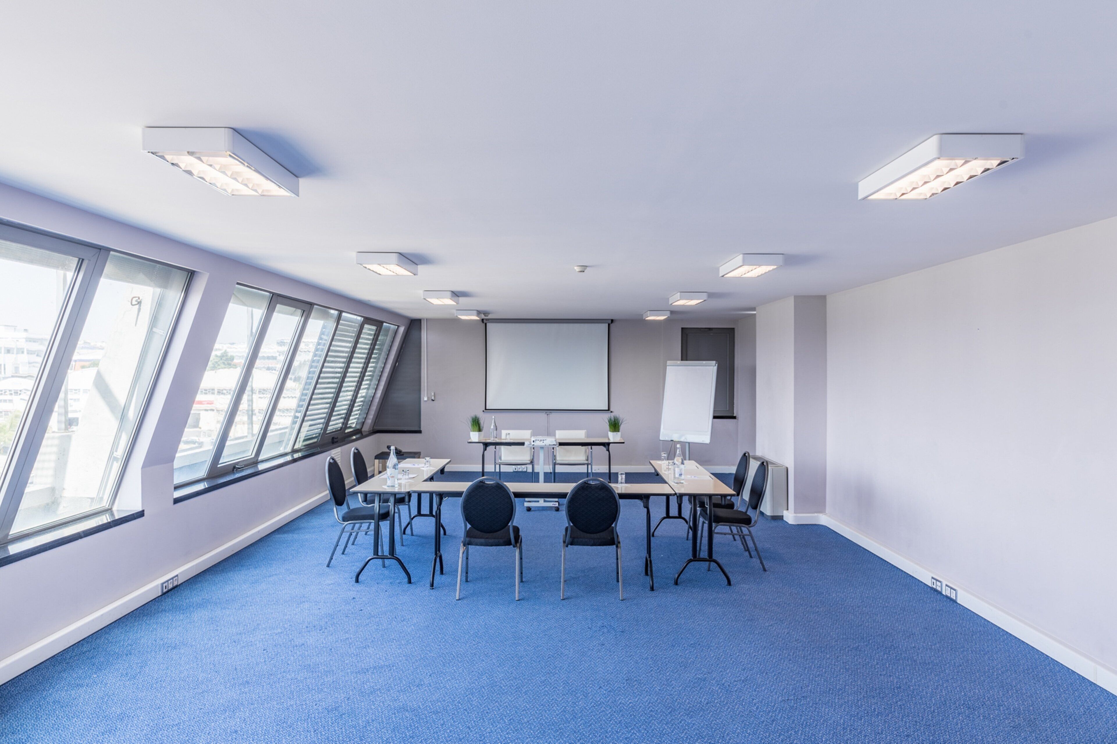 Un salón de conferencias con una mesa larga, sillas, una pantalla de proyector y grandes ventanales.