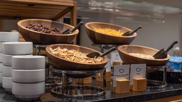 Un bufé con distintos alimentos en recipientes de madera, una estación de café con vasos desechables y un letrero que indica los tipos de comida disponibles.