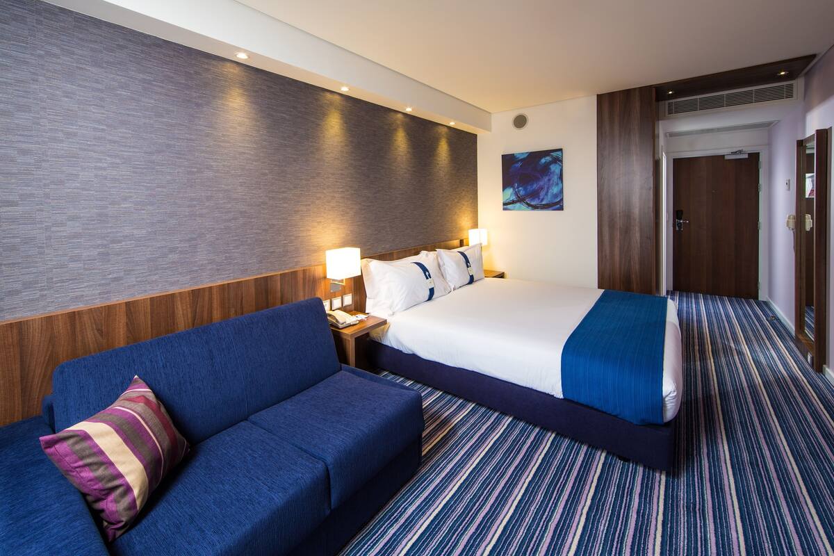 Ett hotellrum med en stor säng, en blå soffa, ett nattduksbord och en väggmonterad lampa.