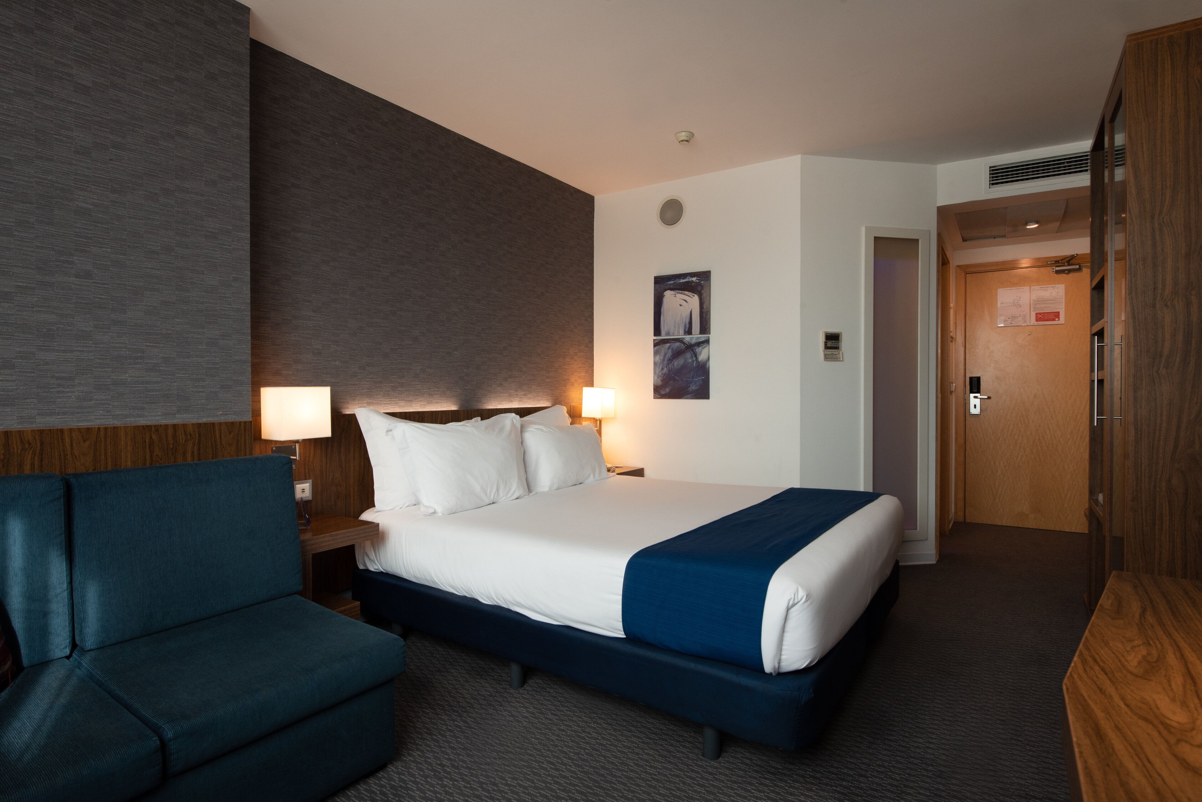 A hotel room with a bed, a desk, a chair, and a large wall with a textured design.