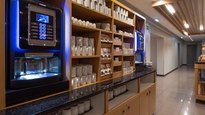 Una estación de café autoservicio con un sistema de pedidos digitales, repisas con tazas y platos, y un interior moderno y limpio.