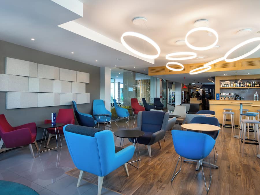 A modern bar area with a variety of seating options including bar stools and chairs, a counter with a selection of beverages, and a ceiling with circular light fixtures.